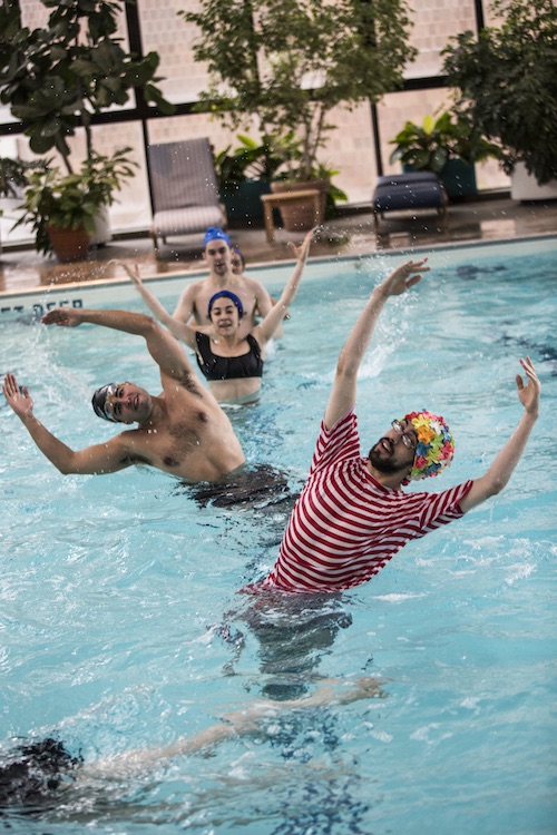 Several performers in bathing suits raise their arms over head while wading in an indoor pool